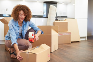 Woman closes a box using a tape dispenser. She is in a room with other boxes. She is moving out of her apartment.